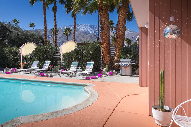 Exterior view of pink walls with a disco ball, patio lounge furniture, and the pool at Desert Dreamer Plum Guide vacation home in Palm Springs, California
