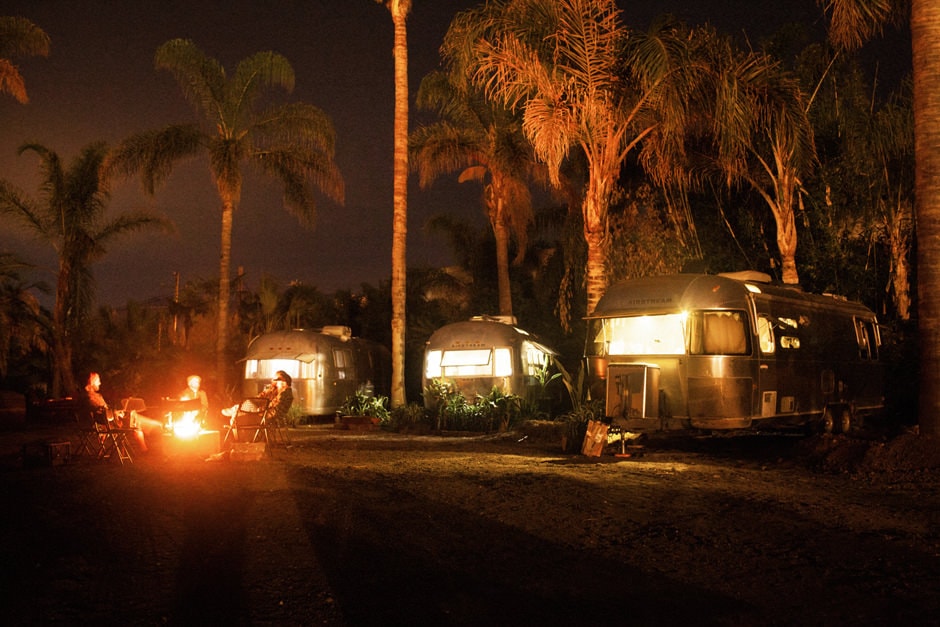Three Airstream trailers at night with a group of people sitting around a fire at Caravan Outpost in Ojai, California.