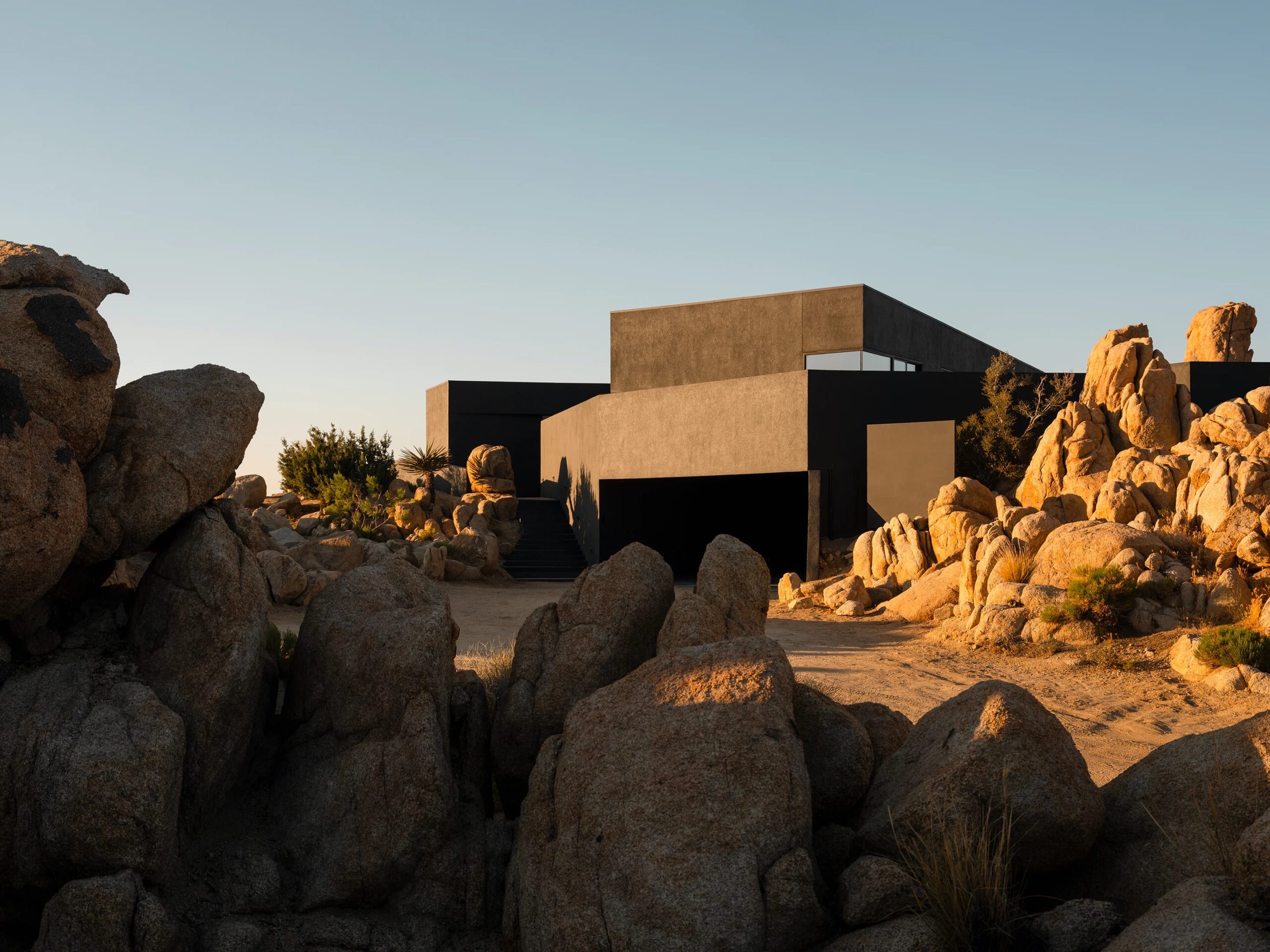 Exterior view of a modern home surrounded by desert landscape and boulders called Black Desert House near Joshua Tree National Park, California.