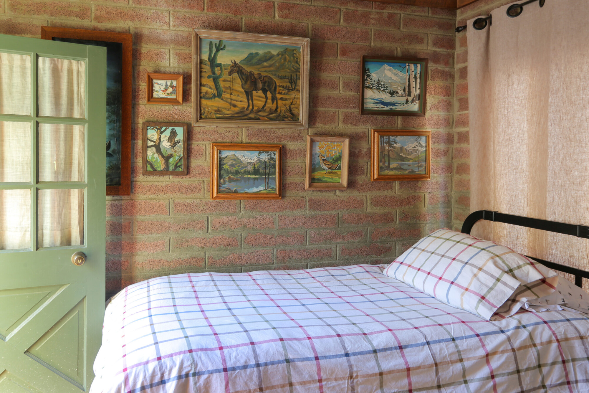 A twin bed set against a brick wall with a collection of paintings in a twin bedroom at Kern River House's River Willow Cabin in Kernville, California.