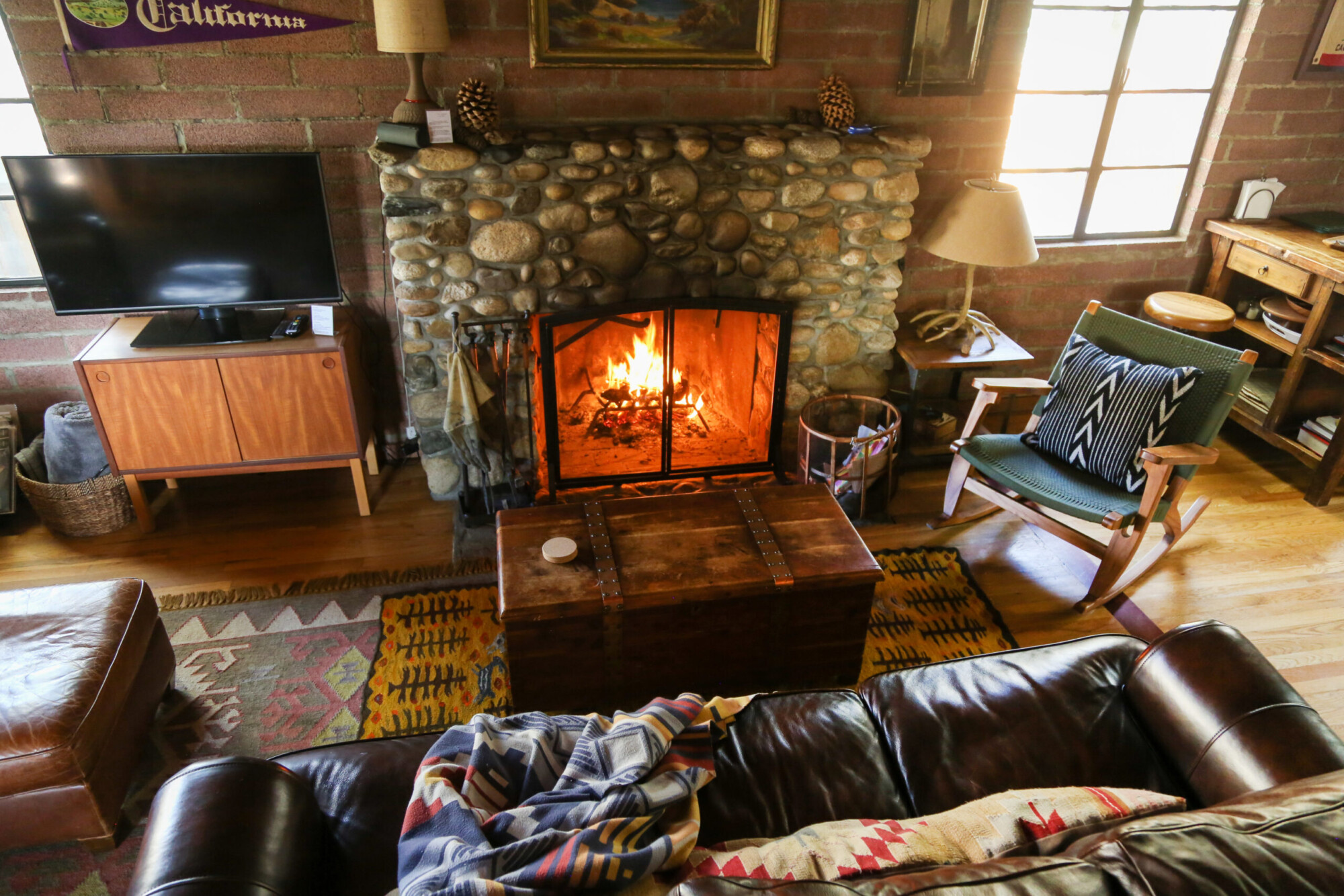View of the cozy living room centered around a fire in a large stone fireplace at Kern River House's River Willow Cabin near Kernville, California.