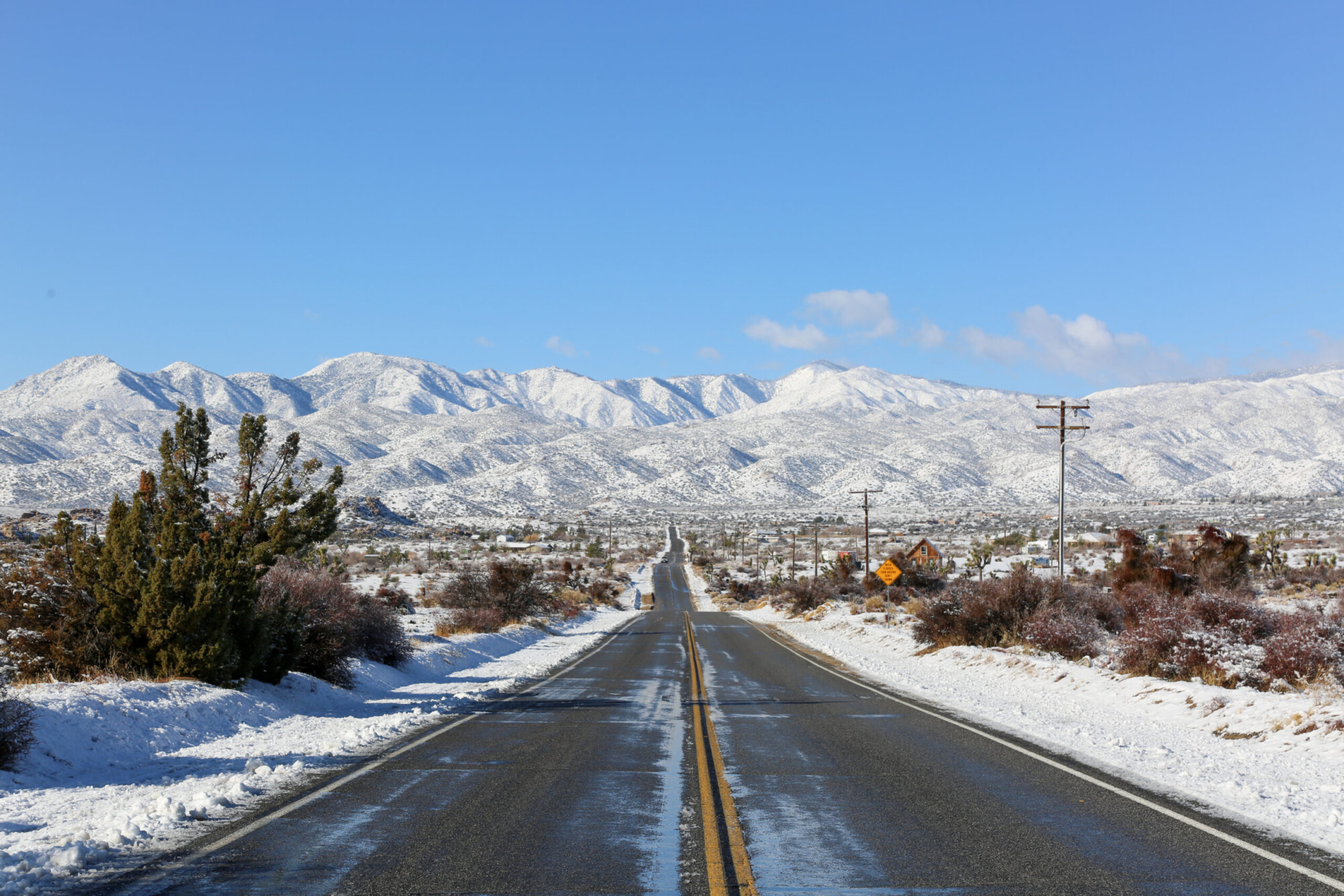 A paved highway flanked in snow with snowy mountains in the background in Pipe's Canyon near Joshua Tree National Park, California.